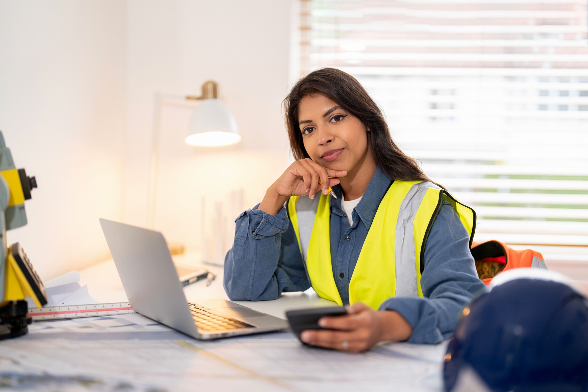 Female land surveyor reviews plans while using smartphone and laptop during office hours at project site